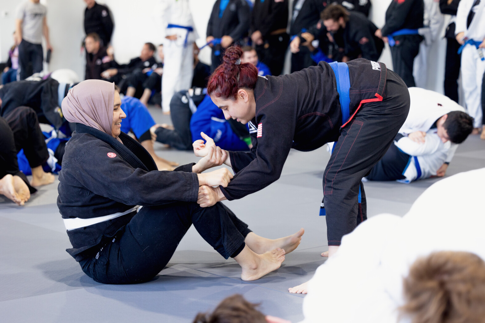 Women training in Brazilian Jiu-Jitsu, one sitting on the ground, the other leaning forward with hands gripping the other's pants